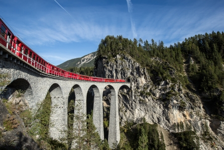 Ein roter Zug der Rhätischen Bahn fährt am Ufer des Flusses durch die beeindruckende Ruinaulta Schlucht in Graubünden. Die Szene wird von hohen, steilen Felswänden in hellen Grau- und Weißtönen dominiert, die sich dramatisch über den Fluss erheben. Einzelne Bäume und grüne Vegetation sind auf den Felsen und am Flussufer zu sehen. Der Himmel ist teilweise blau mit einigen weißen Wolken.