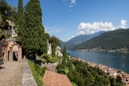 Das malerische Dorf Morcote in der Schweiz, terrassenförmig an einem steilen, bewaldeten Hang am Ufer des Luganersees gelegen. Im Vordergrund führen Wege und Treppen zwischen hohen Zypressen und historischen Gebäuden hinauf zur Kirche Santa Maria del Sasso, deren Turm sichtbar ist. Der weite Luganersee erstreckt sich nach rechts, umgeben von weiteren Bergen unter einem blauen Himmel mit einigen Wolken.